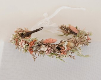 May include: A delicate floral crown made with dried flowers in shades of pink and white. The crown is made with a variety of dried flowers, including small pink flowers, white flowers, and dried grasses. The crown is tied with a white ribbon.