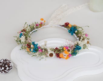 Könnte beinhalten: Ein farbenfroher Blumenkranz mit getrockneten Blumen in Blau, Orange, Rosa und Weiß. Der Kranz ist mit einem weißen Spitzenband verziert und liegt auf einem weißen, wolkenförmigen Tablett.