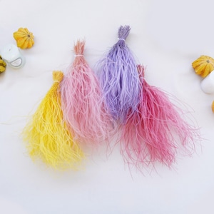 May include: Four bundles of dyed fluffy grass in yellow, pink, purple, and red colors. The grass is arranged in a row on a white surface.