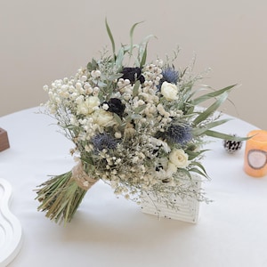 May include: A dried flower bouquet with white gypsophila, cream roses, and blue thistle. The bouquet includes dark blooms and green foliage, tied with twine. A natural and elegant floral arrangement.