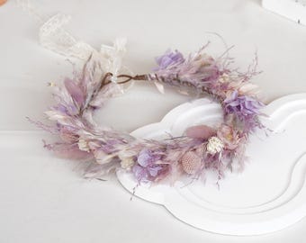 May include: A floral crown made with dried flowers in shades of lavender, pink, and cream. The crown has feathery accents and a brown wire base. It is suitable for weddings or special occasions. A white lace ribbon is attached to the crown.