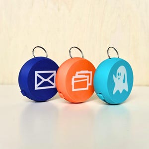 May include: Three circular keyrings in blue, orange, and teal. Each has a white icon: an envelope, files, and a ghost. They have silver rings. The keyrings are likely made of plastic or a similar material.