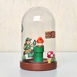 May include: A clear, dome-shaped decorative piece featuring a pixelated video game scene. Inside, a green pipe, a red and white mushroom, and a character are visible. The base is a brown brick design, and the dome is made of clear plastic.