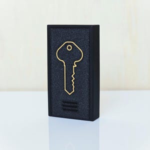 May include: A black rectangular object with a gold key outline on the front. The key design is centered and has a simple, elegant style. Below the key are two horizontal gold lines. The object has a textured surface.