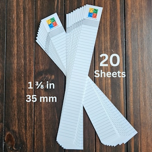 Puede incluir: Una pila de 20 hojas de papel blanco con líneas horizontales, cada una de 35 mm de ancho. La parte superior de cada hoja tiene un logotipo con un diseño colorido y las palabras "OFFICIAL GEOCACHE". Las hojas están dispuestas sobre una superficie de madera oscura.
