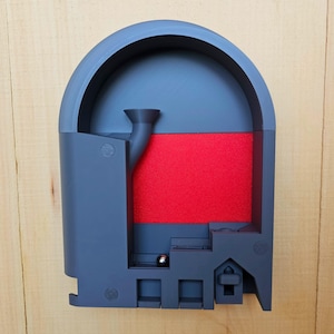 May include: A gray, arched structure with a red rectangular insert and a small metal ball. The structure has a funnel-shaped opening and a series of rectangular cutouts at the bottom. The object is set against a light wood background.