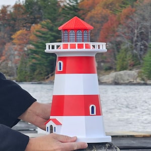 May include: A decorative lighthouse with a red and white striped design. The lighthouse features a red roof and small windows. It has a small door and is held by a person.