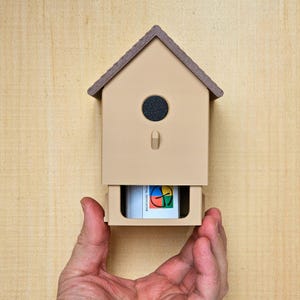 Puede incluir: Una casa para pájaros de color beige con un techo marrón y una abertura circular negra. Se extrae un cajón, revelando un gráfico colorido. La casa para pájaros se sostiene sobre un fondo de vetas de madera clara.