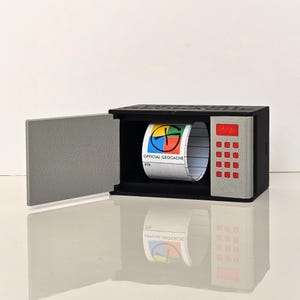 May include: A black miniature safe with a grey door and keypad. Inside, a roll of paper displays the words "OFFICIAL GEOCACHE" and a colourful logo. The safe is designed to hold geocaching logs.