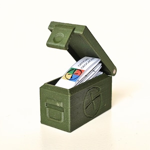 May include: A small, olive green, hinged box with the lid open, revealing a stack of white cards. The cards have the words "OFFICIAL GEOCACHE" printed on them, along with a colorful logo. The box has a cross design on the front.