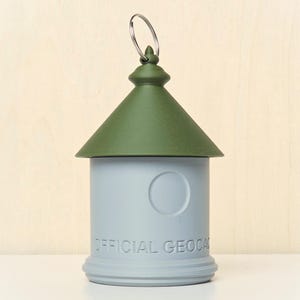 May include: A cylindrical geocache container with a light gray base and a dark olive green conical roof. The base features a circular indentation and the embossed text "OFFICIAL GEOCACHE" along the bottom. A silver metal ring is attached to the top.