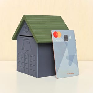 May include: A dark gray house-shaped piggy bank with a green roof, featuring a molded keypad and card slot on the front. Beside it stands a gray Mastercard credit card with the text "world" and "Forward Banking" printed on the surface.