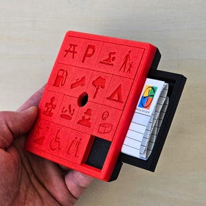 May include: A red rectangular container with various embossed symbols, including a picnic table, a person, and a gas pump. A black drawer slides out, revealing a notepad with the text "TROPICAL GEOCACHE".