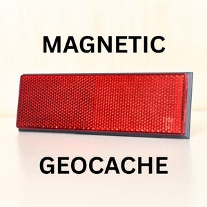 May include: A rectangular red magnetic geocache reflector with a textured, grid-like surface pattern. The object is labeled with the text MAGNETIC above and GEOCACHE below, featuring a dark gray frame surrounding the reflective red center panel.