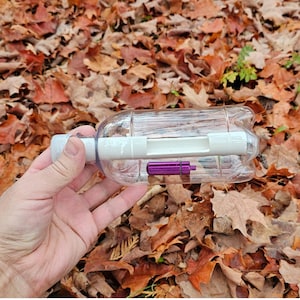 May include: A clear plastic bottle with a white interior and a small, purple cylindrical object inside. The bottle is held in a hand against a backdrop of fallen autumn leaves.