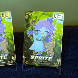 Può includere: Una carta collezionabile rettangolare raffigurante un personaggio dei cartoni animati con capelli viola e un corpo da cervo marrone che abbraccia una creatura blu. La carta ha uno sfondo di foresta verde, dettagli dorati e il testo SPRITE, TEMPORARY, TOKEN UNIT - FAE.