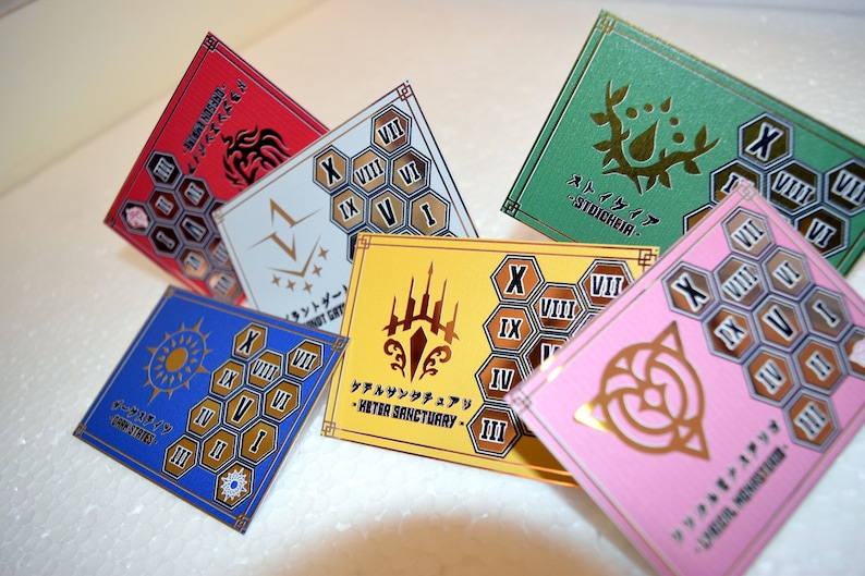Op de afbeelding: Een verzameling kleurrijke, vierkante kaarten met gouden accenten en Japanse tekst. Elke kaart heeft een uniek ontwerp, inclusief symbolen, cijfers en geometrische patronen. De kaarten zijn in tinten rood, blauw, geel, groen en roze.