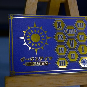 Op de afbeelding: Een blauwe kaart met gouden details, met een zonnesterontwerp en Japanse tekst. De kaart toont ook Romeinse cijfers in gouden zeshoekige vormen. De tekst "DARK STATES" is zichtbaar.