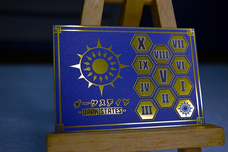 Op de afbeelding: Een blauwe kaart met gouden details, met een zonnesterontwerp en Japanse tekst. De kaart toont ook Romeinse cijfers in gouden zeshoekige vormen. De tekst "DARK STATES" is zichtbaar.