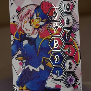 Puede incluir: Una tarjeta plateada con una ilustración colorida de una persona que lleva un sombrero rojo y azul y una chaqueta azul. La tarjeta tiene una cuadrícula hexagonal plateada con los números del 1 al 10.