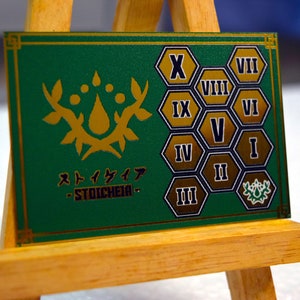 Op de afbeelding: Een groene kaart met gouden accenten en de Japanse tekst "Stoicheia". De kaart heeft een gouden bloemenontwerp en genummerde zeshoekige vormen. De kaart wordt weergegeven op een klein houten ezel.