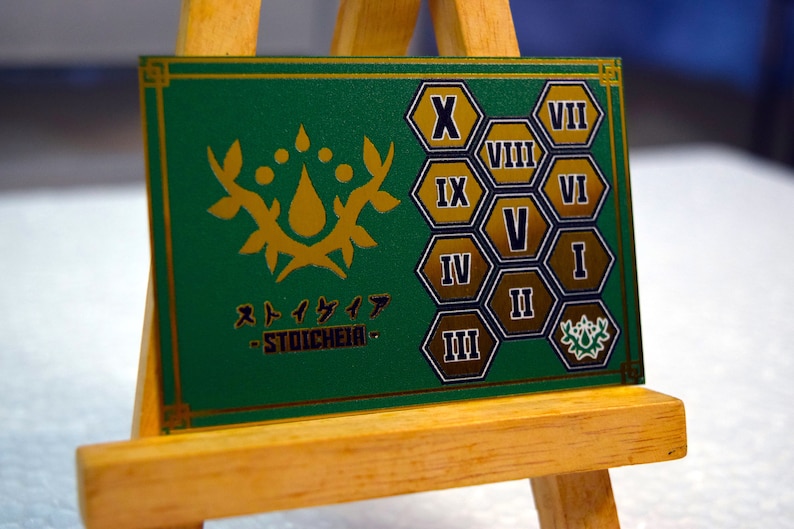 Op de afbeelding: Een groene kaart met gouden accenten en de Japanse tekst "Stoicheia". De kaart heeft een gouden bloemenontwerp en genummerde zeshoekige vormen. De kaart wordt weergegeven op een klein houten ezel.