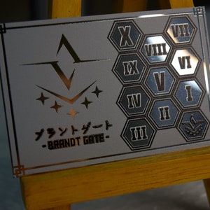Op de afbeelding: Een zilverkleurige rechthoekige plaat met een gouden rand, met een gestileerd embleem en Japanse tekst. De rechterkant toont Romeinse cijfers in zeshoekige vormen. De tekst "BRANDT GATE" is ook zichtbaar.