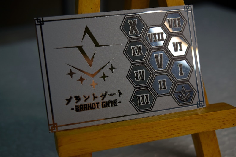 Op de afbeelding: Een zilverkleurige rechthoekige plaat met een gouden rand, met een gestileerd embleem en Japanse tekst. De rechterkant toont Romeinse cijfers in zeshoekige vormen. De tekst "BRANDT GATE" is ook zichtbaar.