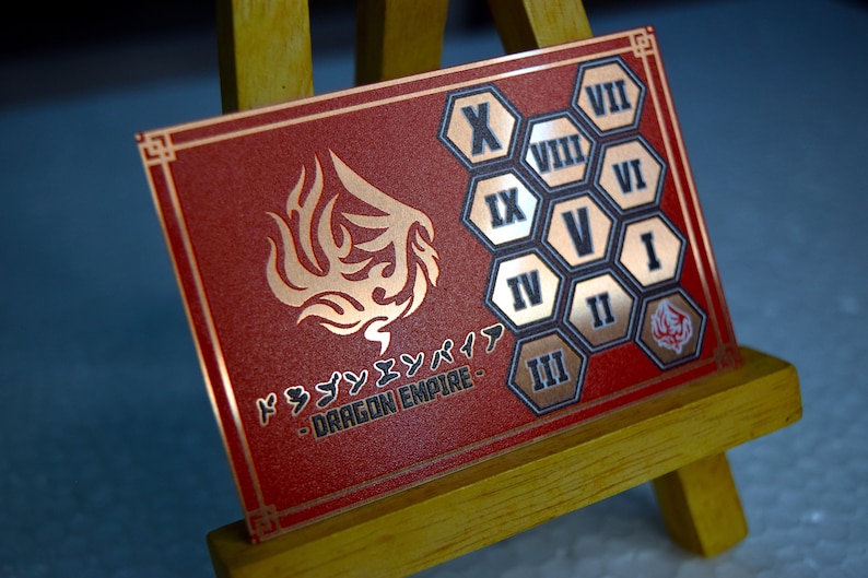 Op de afbeelding: Een rode en gouden Dragon Empire kaart met Japanse tekst en een drakenembleem. De kaart heeft Romeinse cijfers in gouden zeshoekige vormen. De kaart wordt gepresenteerd op een kleine houten ezel.