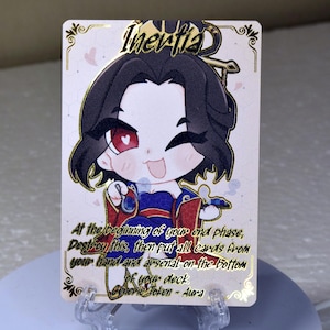 May include: A card game card with a gold border and a cartoon illustration of a girl with black hair and a red and blue dress. The card text reads "Inertia" and "At the beginning of your end phase, Destroy this, then put all cards from your hand and arsenal on the bottom of your deck. Generic Token - Aura".