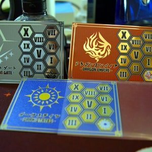 Op de afbeelding: Drie rechthoekige kaarten met Japanse tekst en geometrische ontwerpen. E&eacute;n is zilver met een wit logo en de tekst "BRANDT GATE". Een andere is rood met een gouden drakenontwerp en de tekst "DRAGON EMPIRE". De derde is blauw met een zonontwerp.