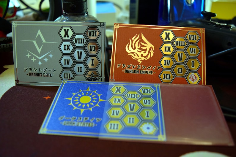 Op de afbeelding: Drie rechthoekige kaarten met Japanse tekst en geometrische ontwerpen. E&eacute;n is zilver met een wit logo en de tekst "BRANDT GATE". Een andere is rood met een gouden drakenontwerp en de tekst "DRAGON EMPIRE". De derde is blauw met een zonontwerp.