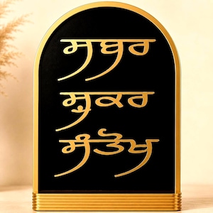 May include: An arched sign with a black background and a gold frame. The sign features three lines of gold text in Gurmukhi script. The base of the sign is rectangular with horizontal gold ridges, providing a stable foundation for the upright display.