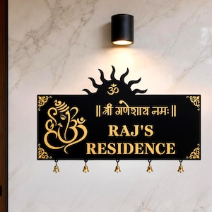 Peut inclure: Une plaque murale rectangulaire noire avec des inscriptions dorées "RAJ'S RESIDENCE" et "॥ श्री गणेशाय नमः ॥". Elle présente une icône dorée de Ganesh, un symbole Om, une bordure supérieure en forme de soleil et cinq petites clochettes en laiton.