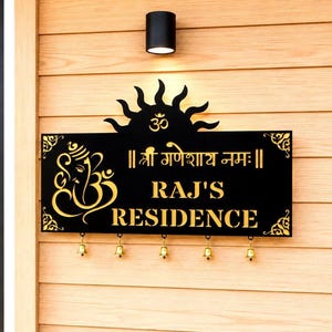 Peut inclure: Une plaque de maison rectangulaire noire et dorée avec un motif de soleil et un symbole Om au sommet. La plaque affiche le texte "RAJ'S RESIDENCE" et "॥ श्री गणेशाय नमः ॥" avec un motif Ganesh. Cinq petites cloches en laiton pendent en bas.