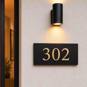 May include: A rectangular black house number plaque featuring the gold-colored digits 302, mounted on a light-colored wall beneath a cylindrical black wall-mounted light fixture that casts a warm glow downward onto the sign.