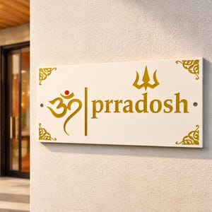 May include: A rectangular white sign with gold lettering and decorative corner accents. The sign features the word "prradosh" in gold, along with a symbol and a red dot. The sign is mounted on a textured white wall.