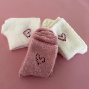 Pode incluir: Três pares de meias felpudas com corações bordados. Um par é rosa e dois pares são brancos.