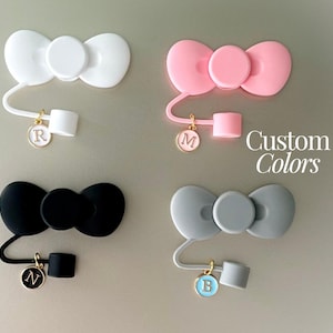 May include: Four different colored silicone bow-shaped cable organizers with a small charm attached to each. The charms are gold with a letter on them. The colors of the bows are white, pink, black, and gray. The text "Custom Colors" is visible in the image.