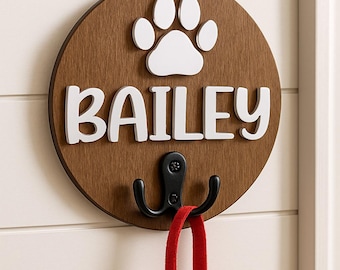 May include: A circular brown wooden wall hook featuring a white paw print icon and the name BAILEY in white raised lettering. A black metal double hook is attached to the center, holding a red leash with a silver metal clasp at the bottom.