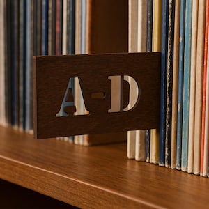 May include: A brown wooden record divider, featuring the cut-out letters "A-D". It's positioned on a wooden shelf, separating a collection of vinyl records with colourful spines.
