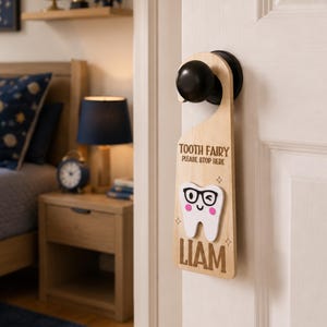 Op de afbeelding: Houten deurhanger met een tandfee-thema. Het lichtgekleurde houten bord heeft de tekst "TOOTH FAIRY PLEASE STOP HERE" en een lachende tandafbeelding met een bril. De naam "LIAM" staat eronder gedrukt.