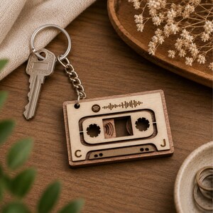 May include: A wooden cassette tape keyring with a silver key and chain. The keyring features a Spotify logo and a sound wave design. The letters "C" and "J" are visible on the keyring. The keyring is made of light-coloured wood.