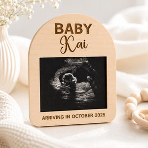 May include: A light-coloured wooden frame with an arched top, displaying a sonogram image. The frame is engraved with the words "BABY Kai" and "ARRIVING IN OCTOBER 2025". The frame is made of light-coloured wood.