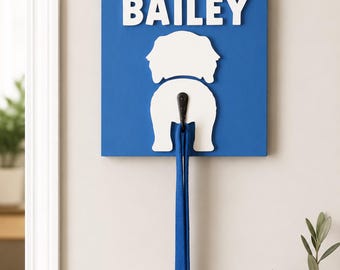 May include: A square blue wall-mounted hook featuring the white text BAILEY above a white silhouette of a dog's rear end. A black metal hook is attached to the centre of the dog shape, holding a long blue lead hanging straight down.