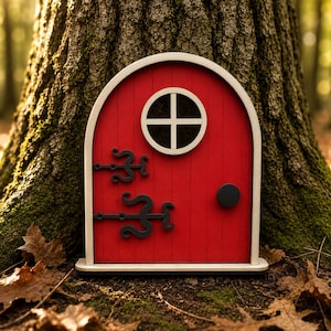 May include: A miniature red fairy door with a white arched frame and a circular window with a cross pattern. It has black decorative hinges and a black doorknob. The door is placed against a tree trunk.