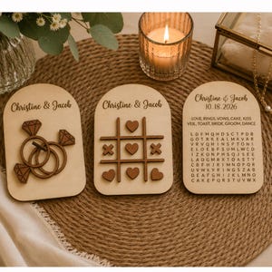 May include: Three arched wooden signs featuring the names Christine & Jacob. The first sign displays two interlocking rings with diamond accents. The second sign shows a tic-tac-toe board with hearts and X's. The third sign features a word search puzzle.