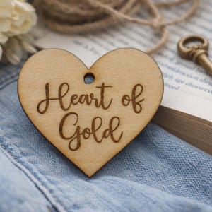 May include: A wooden heart-shaped ornament with the phrase "Heart of Gold" engraved in a cursive font. The ornament has a small hole at the top for hanging. The background includes a book, twine, and a key.