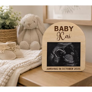 May include: A wooden frame with the words "BABY Kai" engraved at the top and a sonogram image in the centre. The text "ARRIVING IN OCTOBER 2025" is at the bottom. A stuffed bunny, baby shoes, and a blanket are in the background.