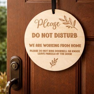 May include: A circular wooden sign with the text "Please DO NOT DISTURB". It also states "WE ARE WORKING FROM HOME" and asks for parcels to be left at the door. The sign is suspended by a black ribbon and is attached to a wooden door.